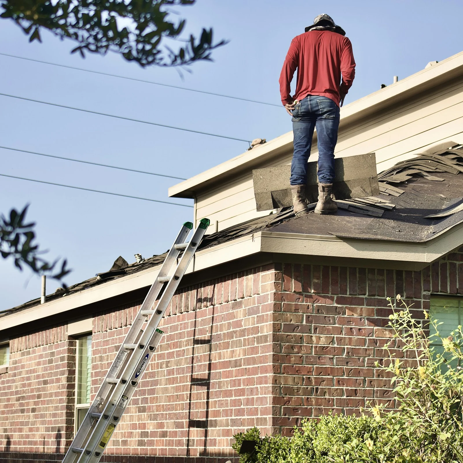 A roofer standing on the roof of a brick suburban home, a ladder against the wall below